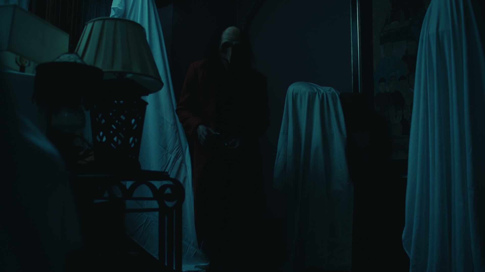 Dark room with covered objects and a person standing in the background, dimly lit, with blue tones.