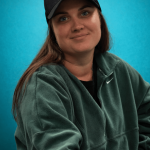 Young woman wearing a black cap and green Nike sweatshirt, smiling against a blue background.