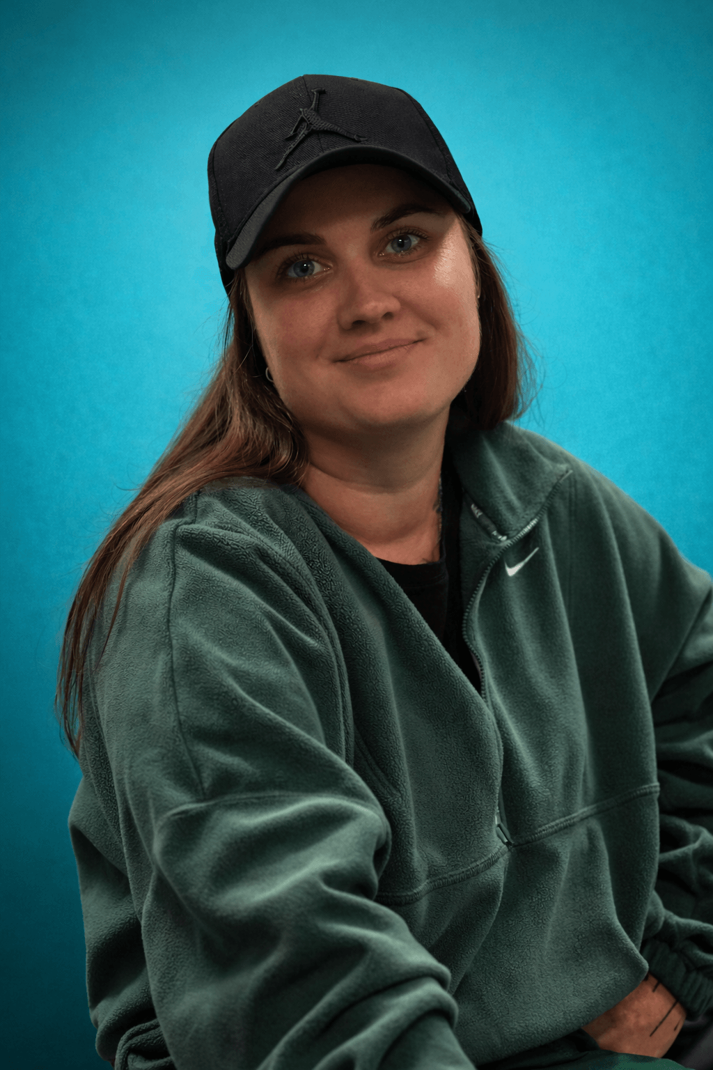 Young woman wearing a black cap and green Nike sweatshirt, smiling against a blue background.