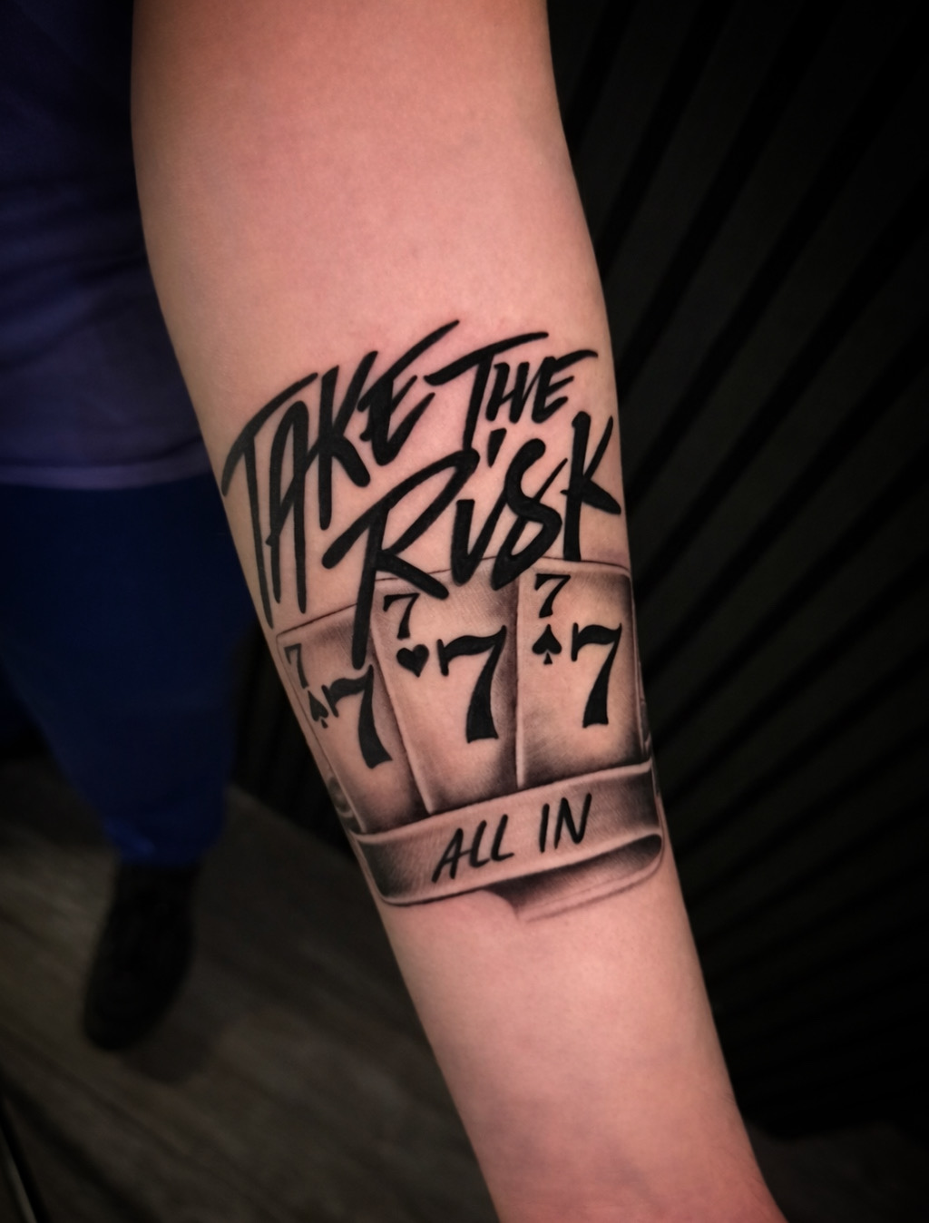 Tattoo on person's arm with text 'Take the risk' and 'All in', and numbers '7-7-7'.