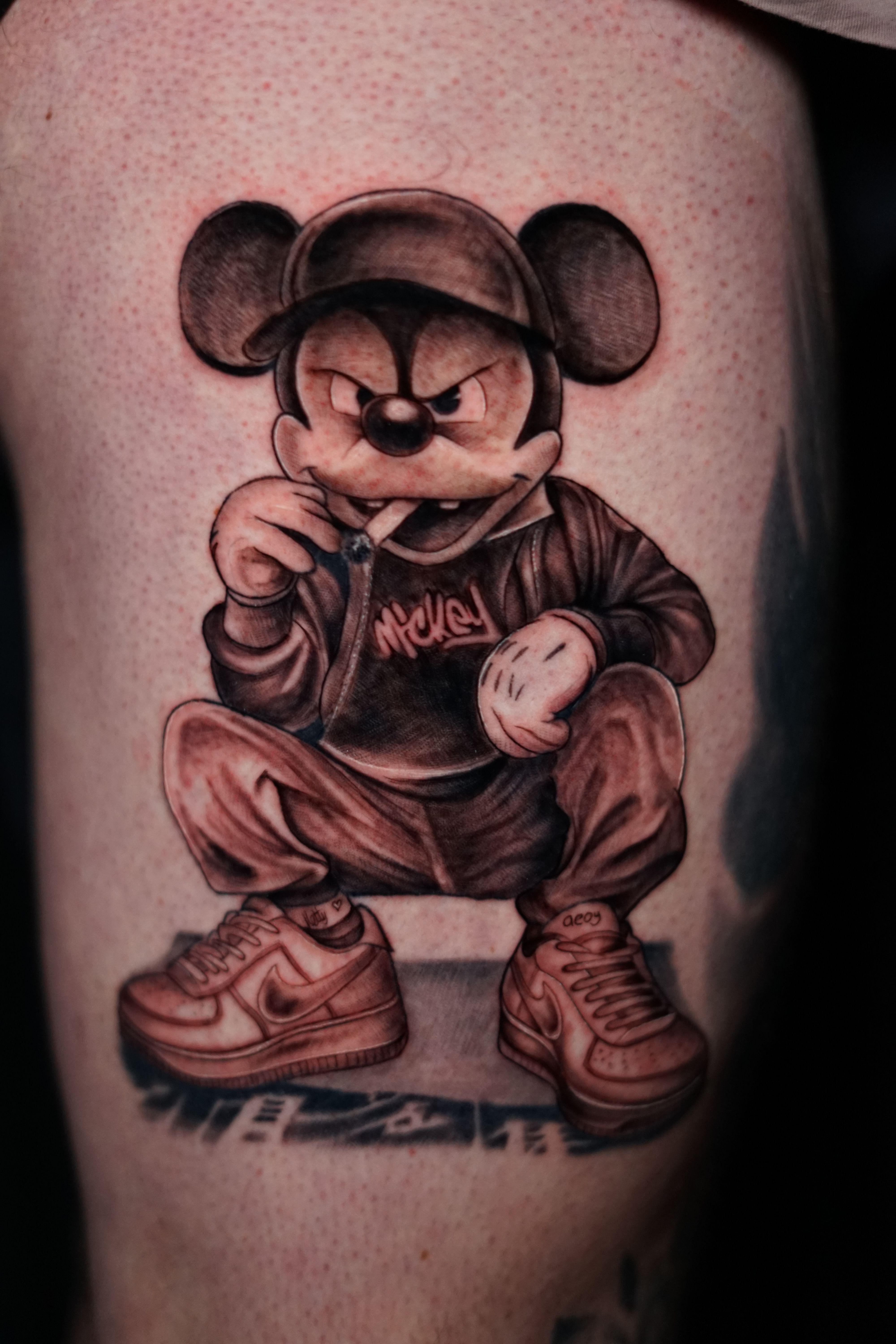Tattoo of a cartoon character with a skull face, wearing sunglasses, a hoodie, and sneakers, holding a small object.