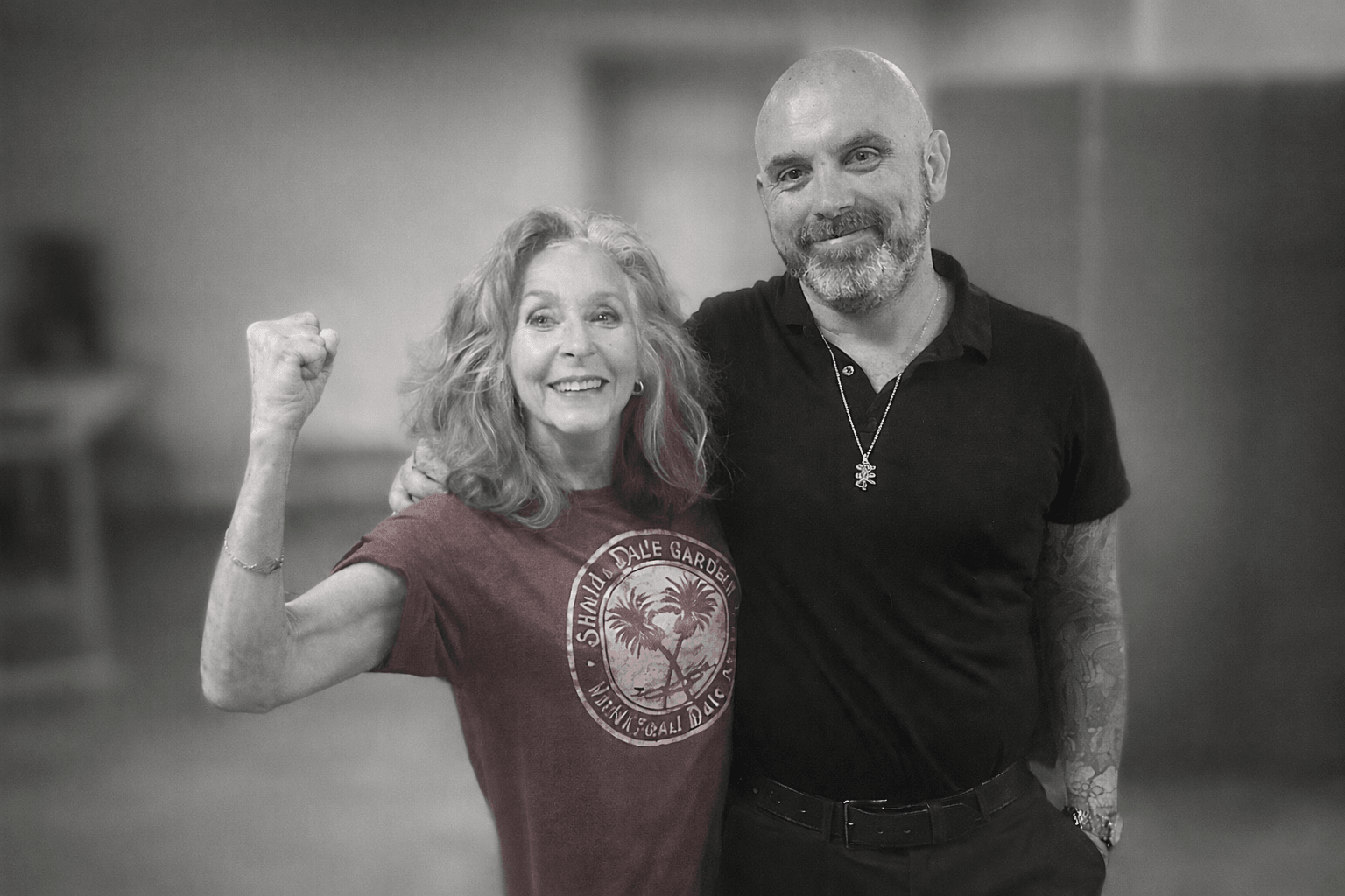 Two people smiling, one woman raising her fist, the other man standing beside her, both in casual clothing, indoors.