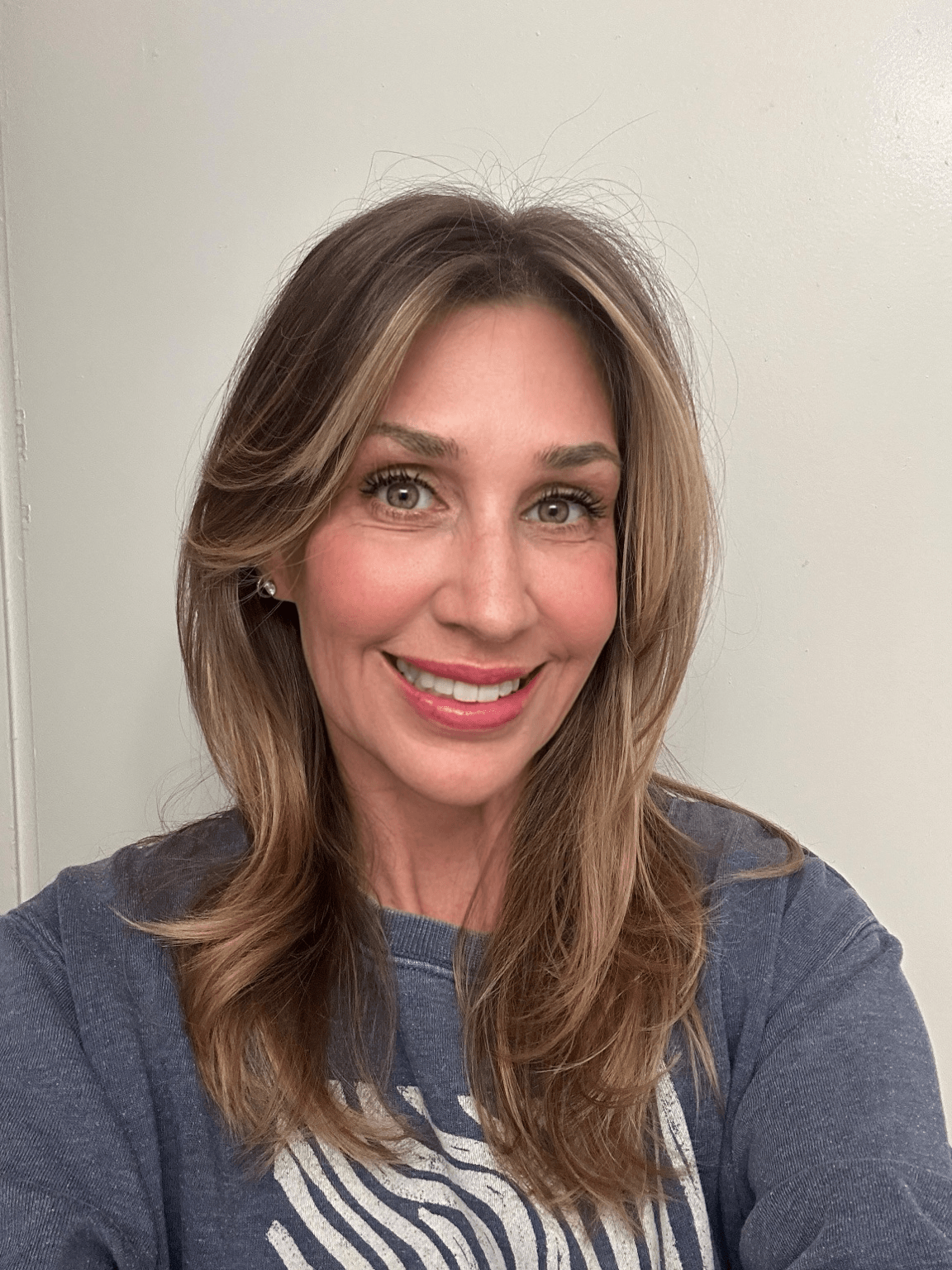 Woman with long, wavy hair smiling, wearing a gray shirt, standing against a plain light-colored wall.