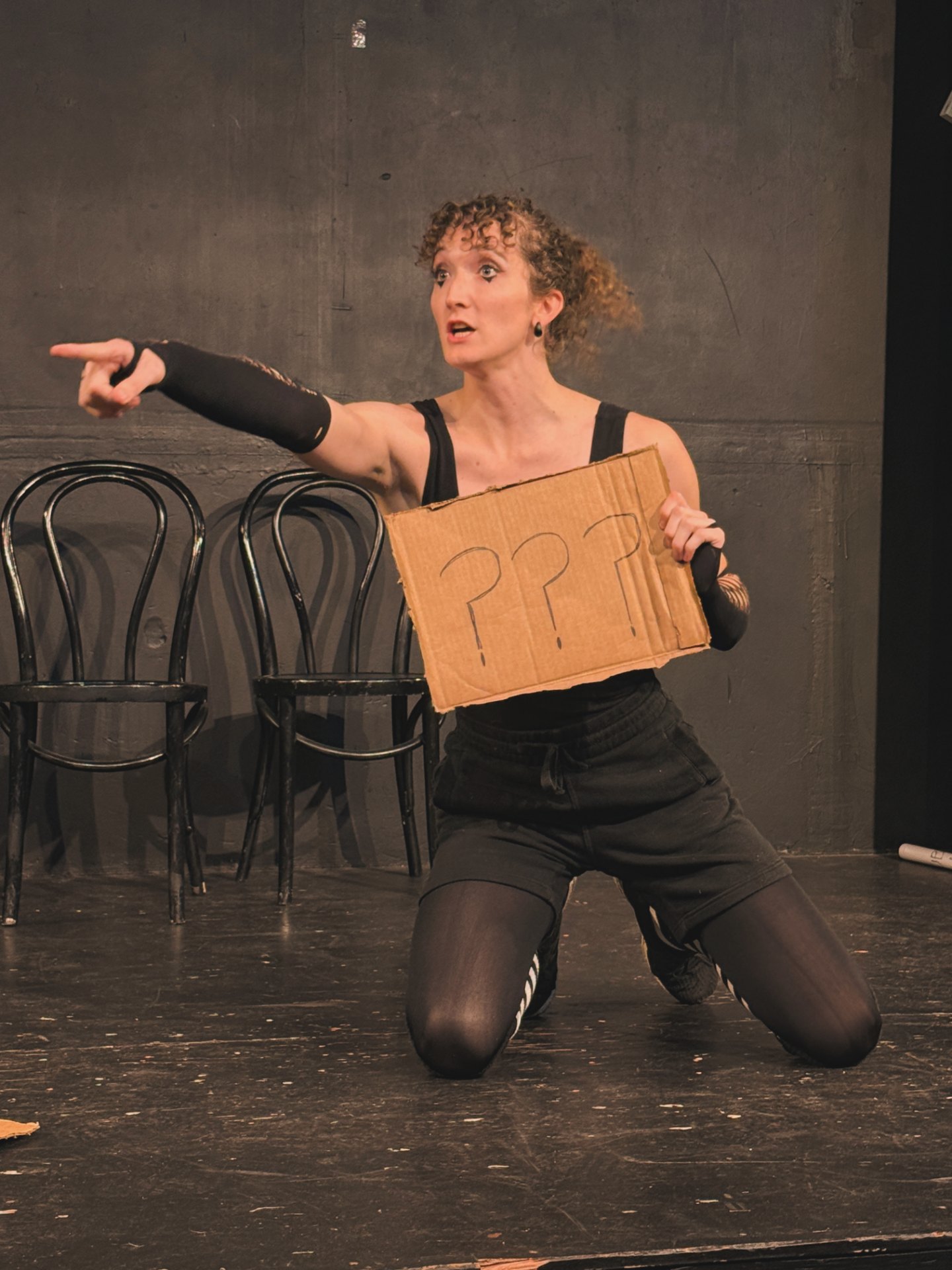 Performer kneeling on stage holding a cardboard sign with question marks, pointing to the left, with chairs in background.