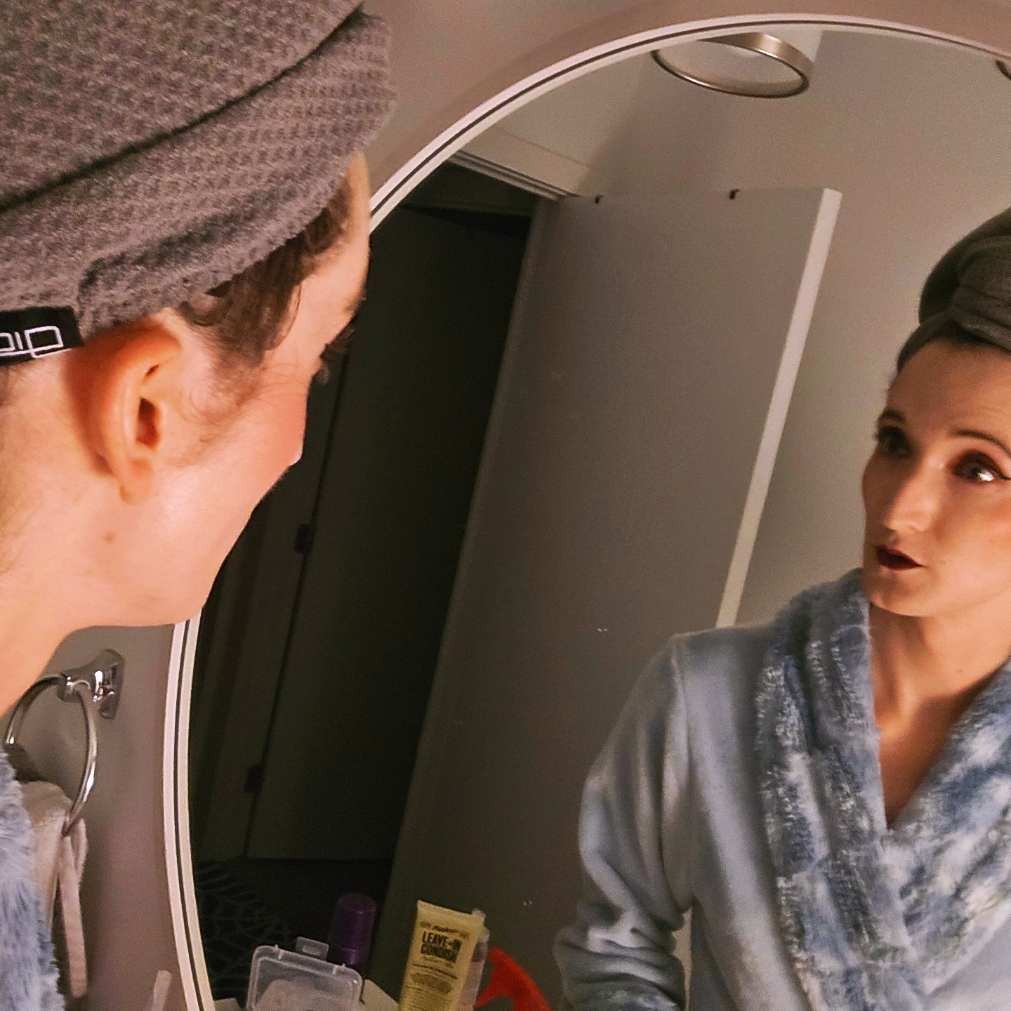 Woman looking at her reflection in a mirror, wearing a gray robe and a head covering, in a room with a door and a mirror.