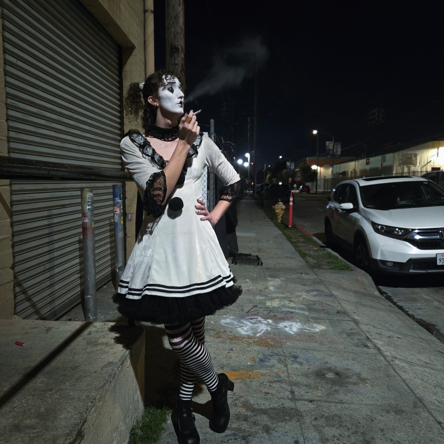 Person dressed as a mime with makeup, wearing a white dress with black accents, standing on a sidewalk at night, smoking.