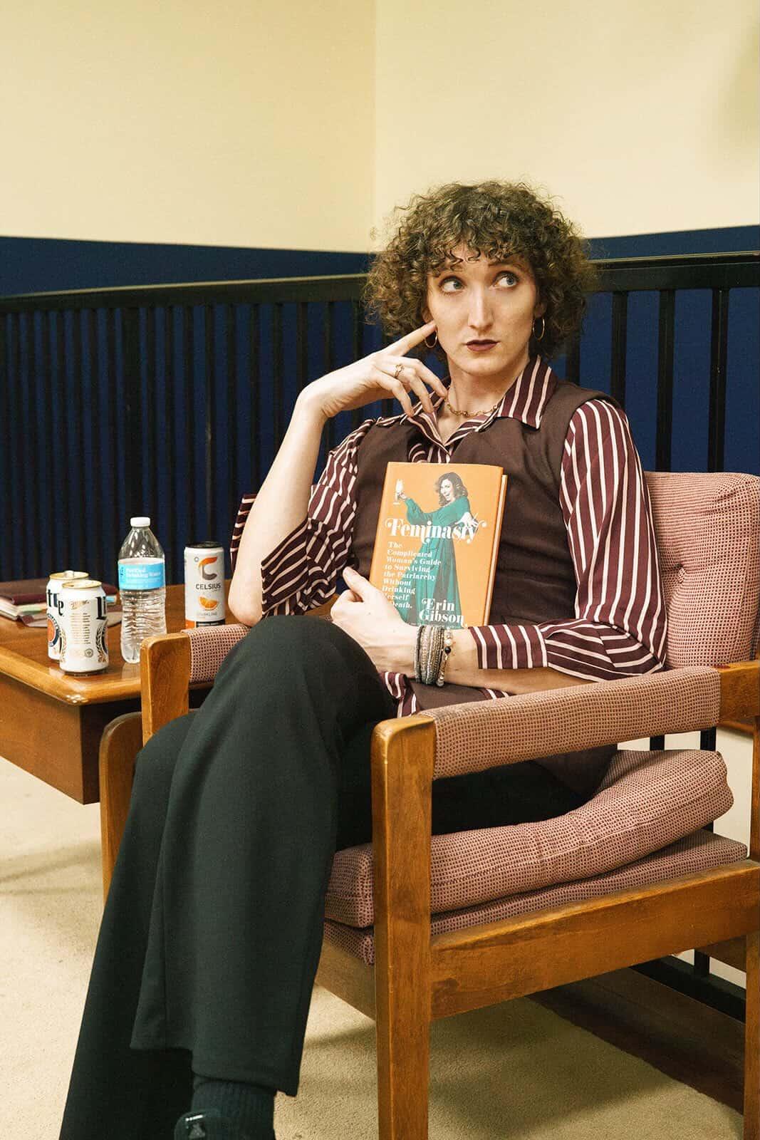 Person with curly hair sitting on a chair, holding a book, with a thoughtful expression, in a room with a blue wall.