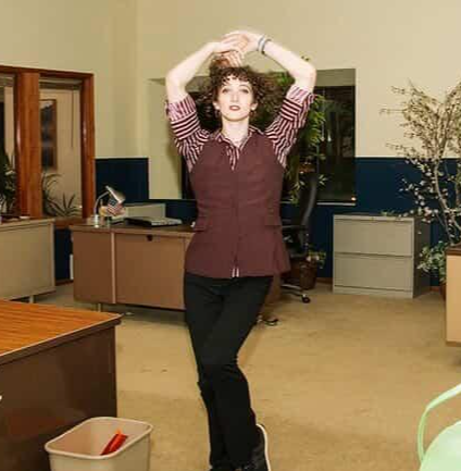 Woman with curly hair standing in an office, arms raised above head, wearing a striped shirt and dark pants.