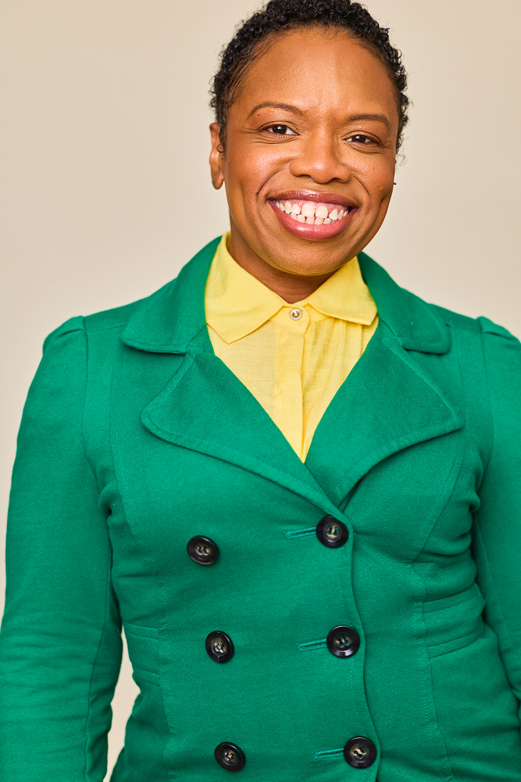 Smiling woman with short curly hair wearing a green blazer and yellow shirt against a plain background.
