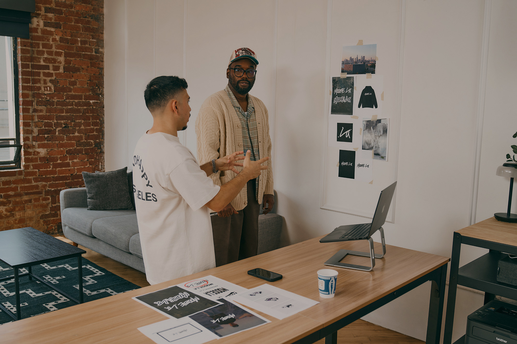Two men talking in a modern office with a desk, laptop, and wall photos, one gesturing with hands, the other listening.