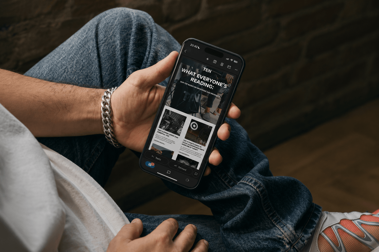 Person holding a smartphone displaying a news app, sitting with legs crossed, wearing jeans and a bracelet, against a brick wall background.