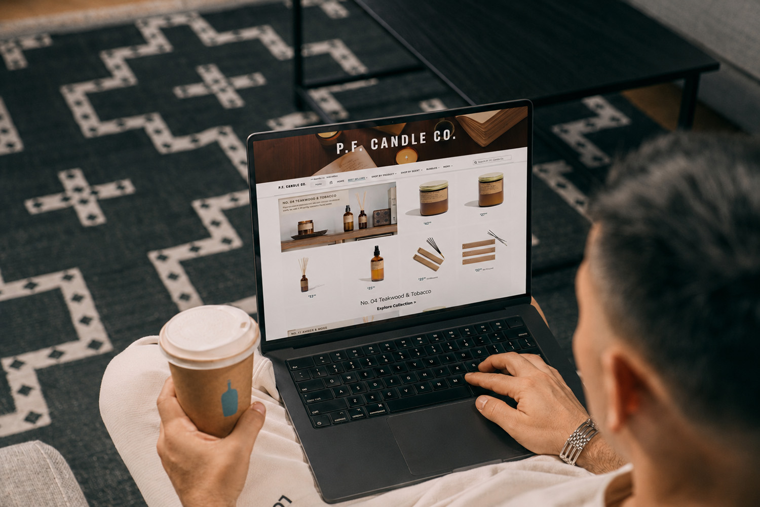 Person holding a coffee cup looking at a laptop displaying a candle company's website, in a room with patterned carpet.