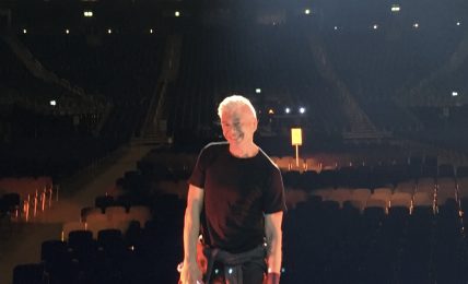 Person with light-colored hair standing on stage in an empty theater, facing away from the camera, with rows of seats behind.