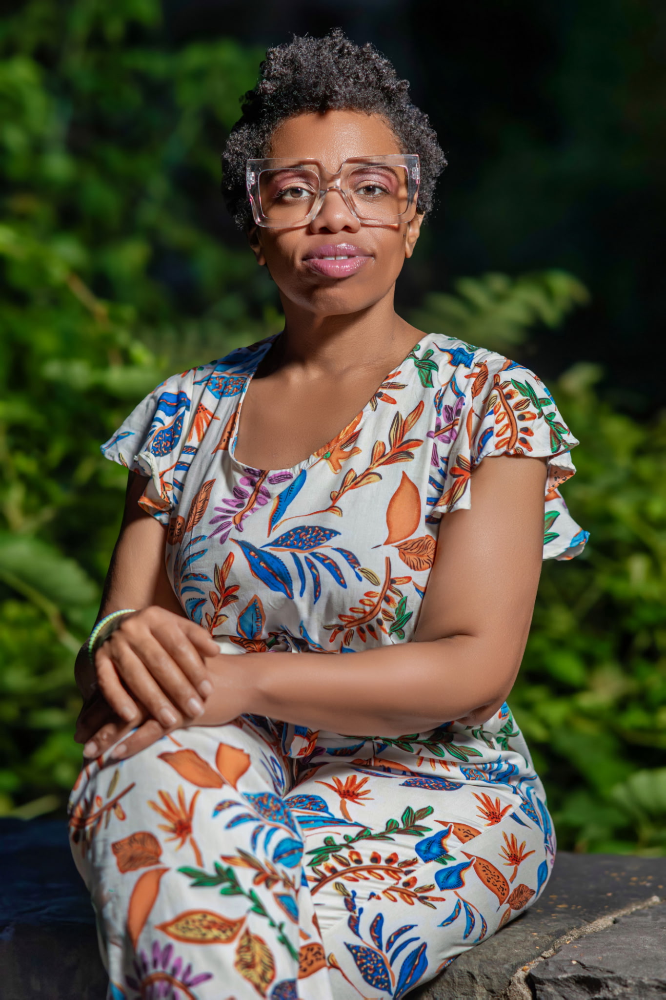 Young woman with glasses and curly hair sitting outdoors, wearing a colorful floral dress, with greenery in background.