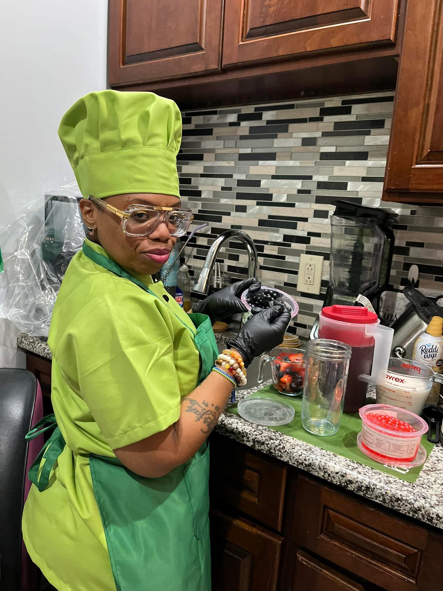 Person in green chef outfit and hat preparing food in kitchen, wearing glasses and black gloves, with various containers on counter.