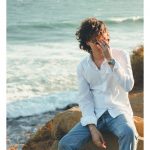 Person with curly hair wearing a white shirt and jeans sitting on rocks near the ocean, covering face with hand.
