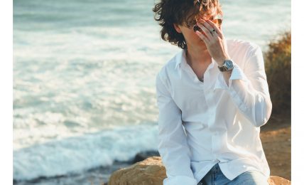 Person with curly hair wearing a white shirt and jeans sitting on rocks near the ocean, covering face with hand.