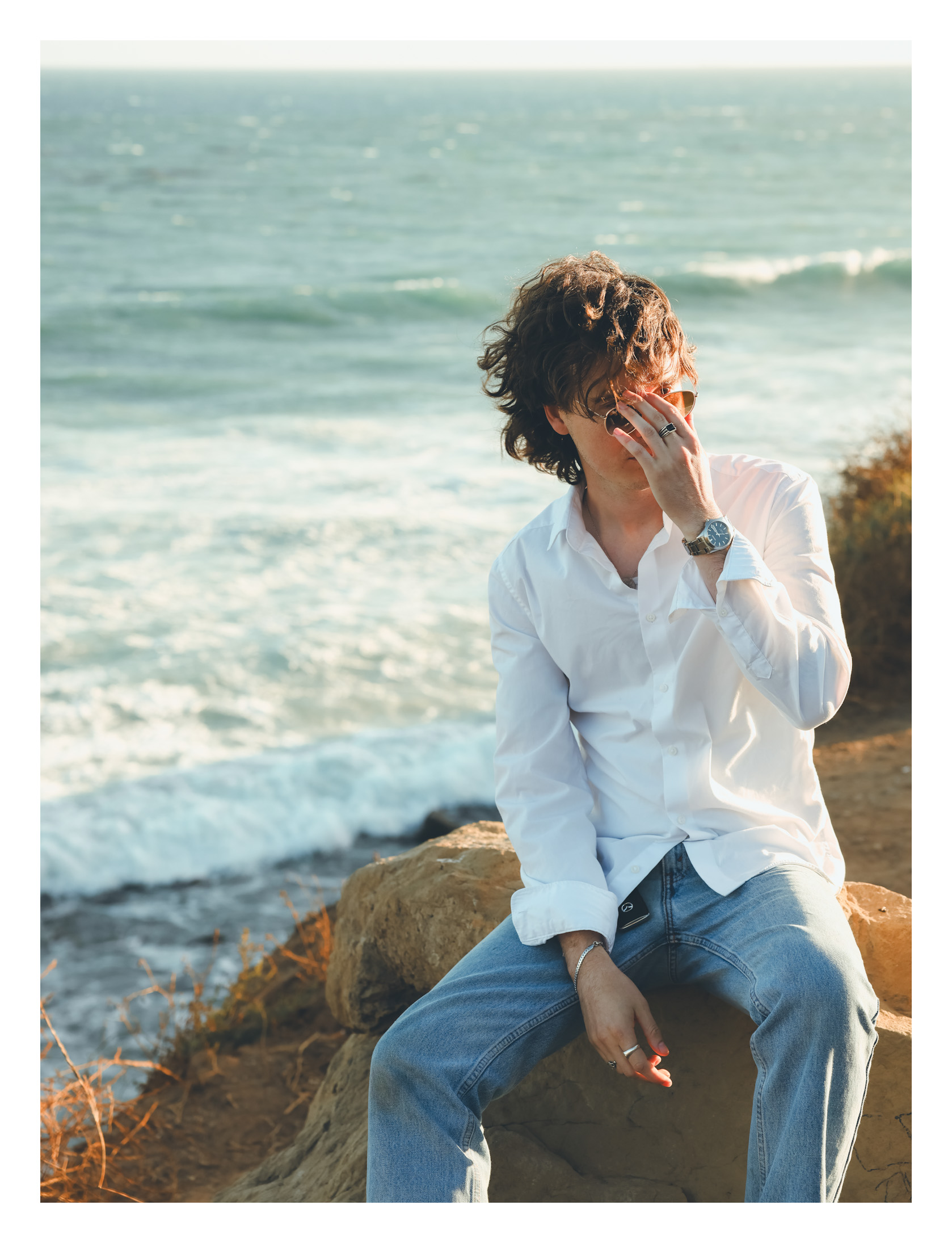 Person with curly hair wearing a white shirt and jeans sitting on rocks near the ocean, covering face with hand.