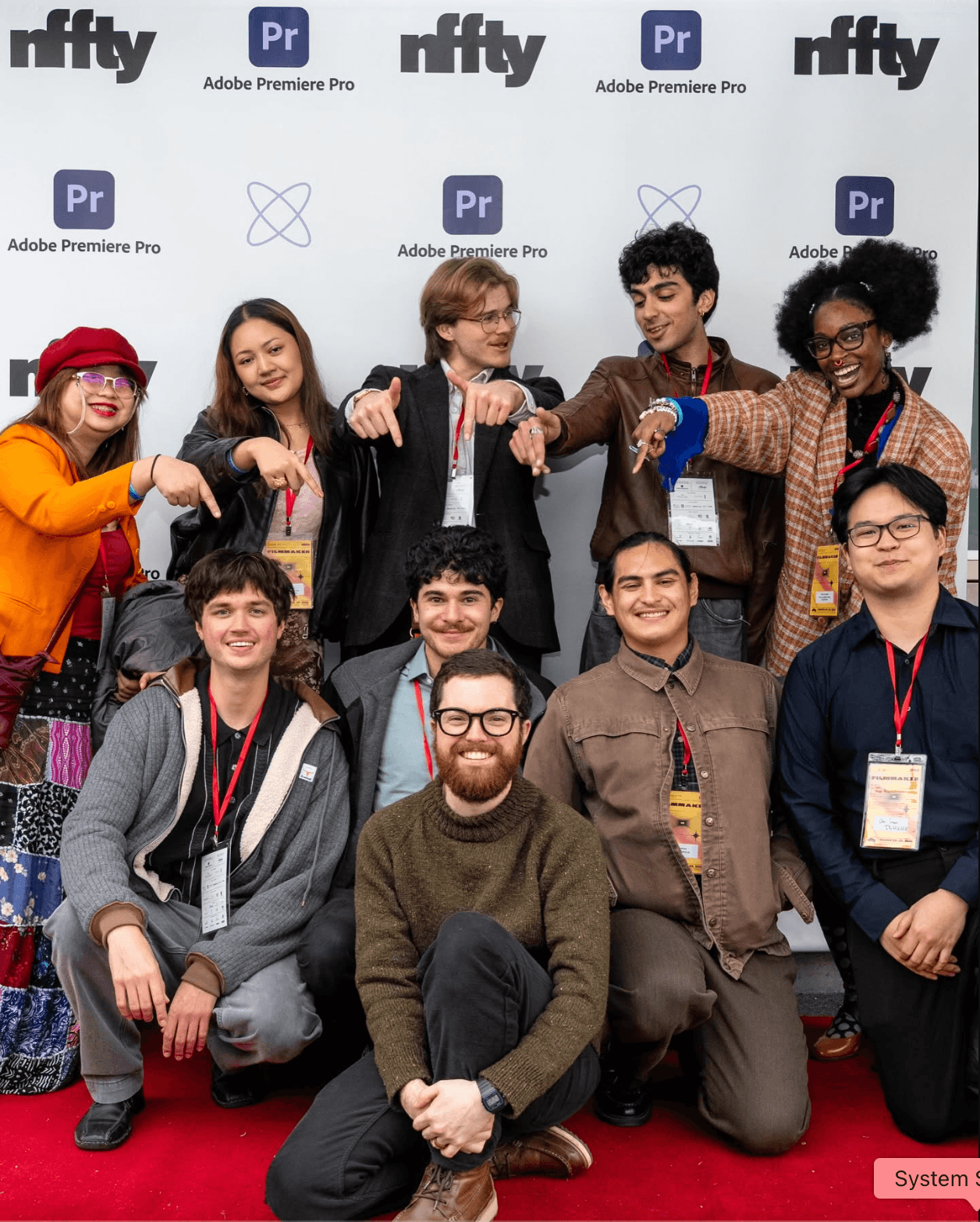 Group of ten diverse people smiling and pointing at camera, standing on red carpet, background with logos, some wearing badges.