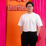 Young man with glasses in white t-shirt and black pants standing in front of colorful backdrop with text.