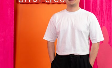 Young man with glasses in white t-shirt and black pants standing in front of colorful backdrop with text.