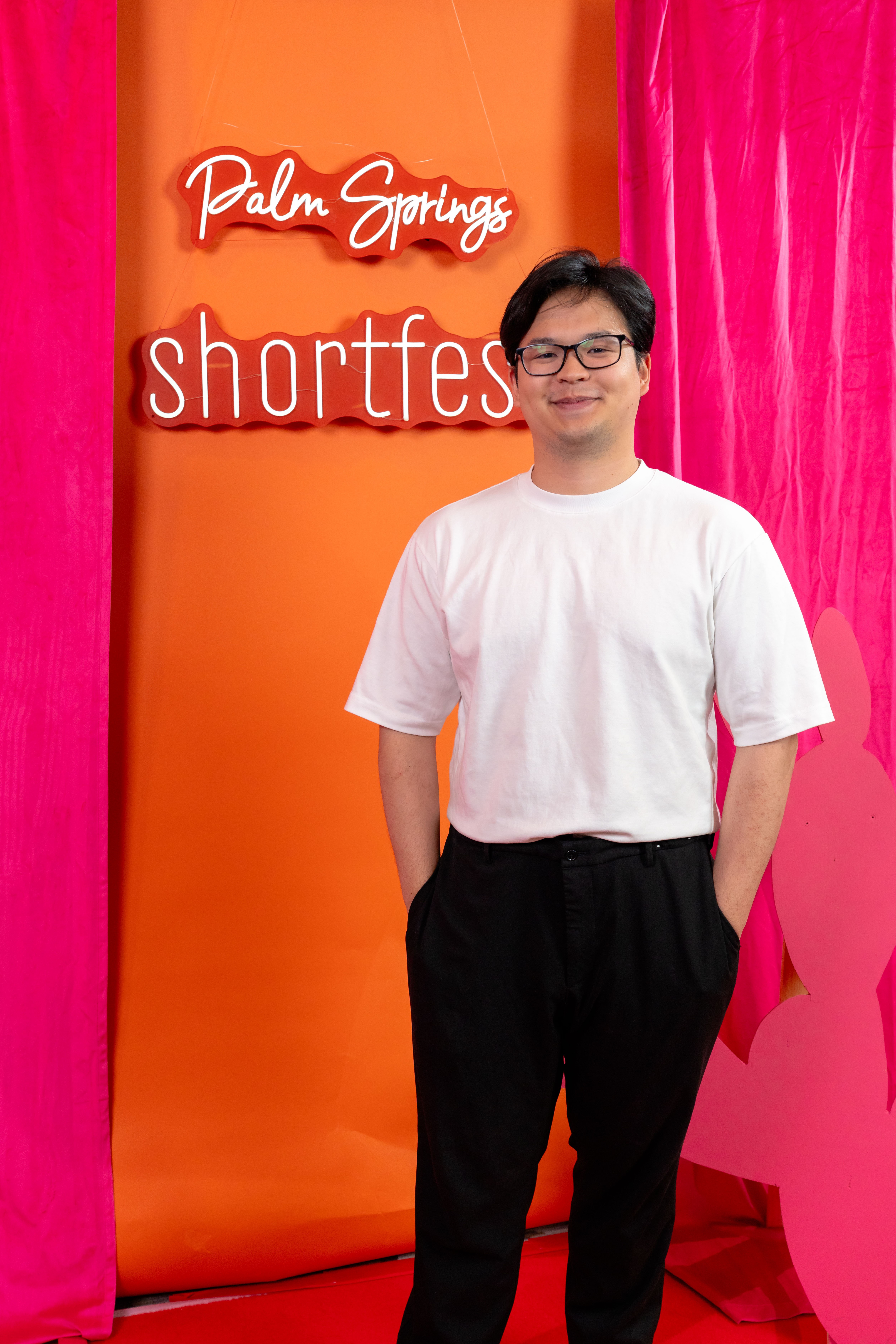 Young man with glasses in white t-shirt and black pants standing in front of colorful backdrop with text.