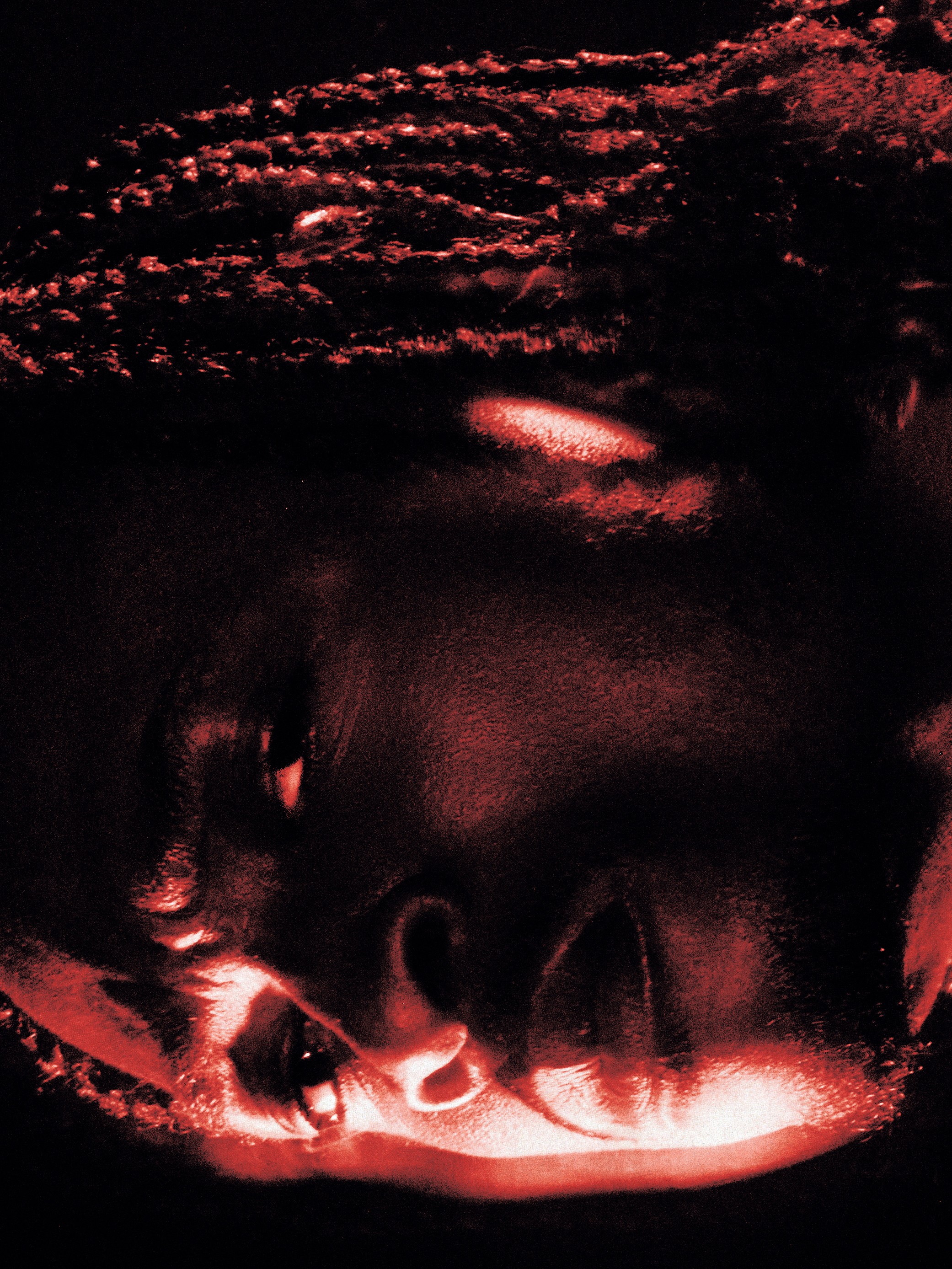 Close-up of a person's face illuminated by red light, with eyes closed and head tilted sideways.