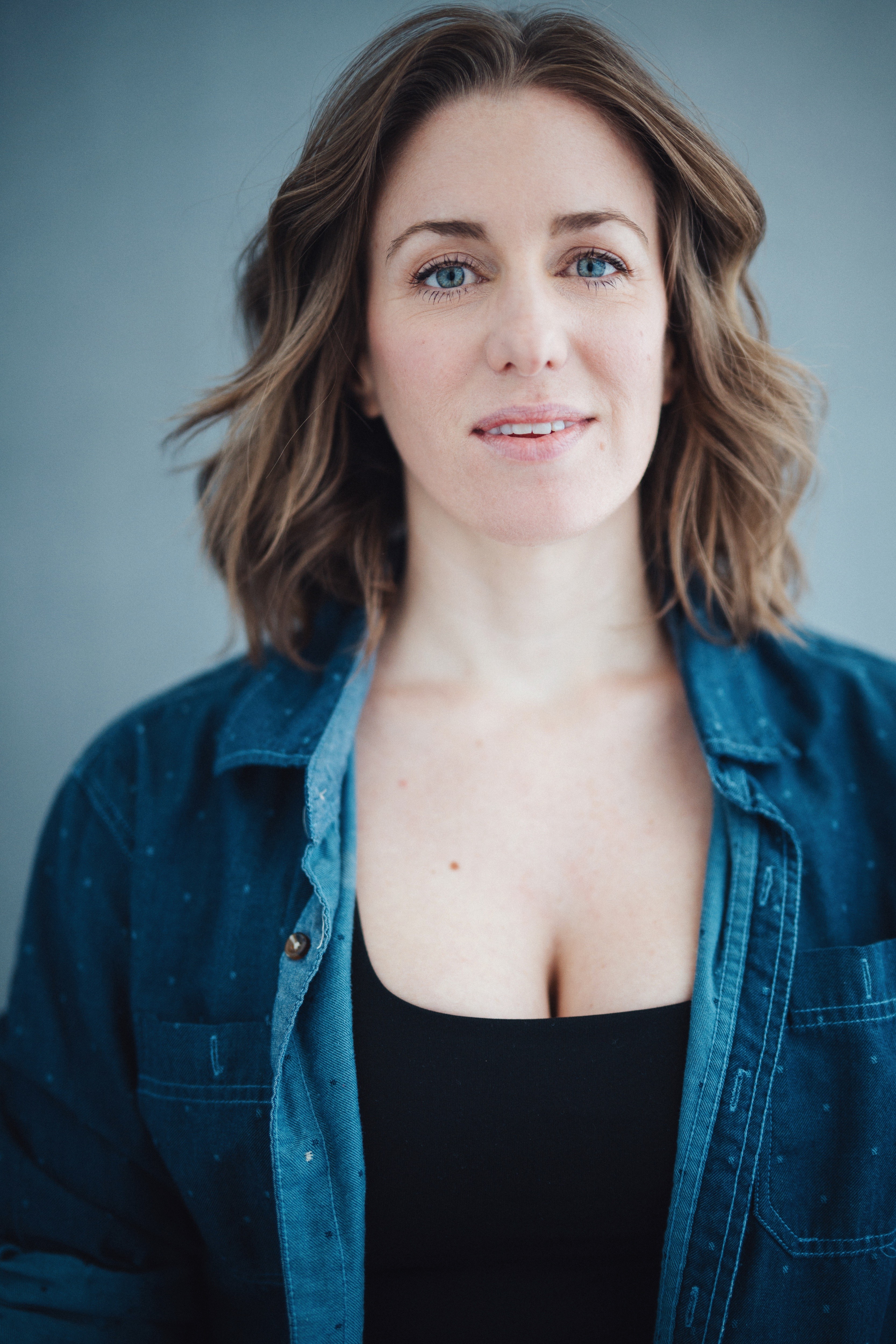 Portrait of a woman with shoulder-length wavy brown hair, wearing a denim shirt over a black top, against a plain background.