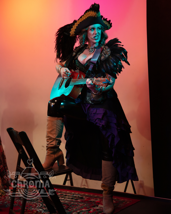 Person dressed as a pirate with a hat, feathered accessories, and a guitar, standing on a stage with a colorful background.