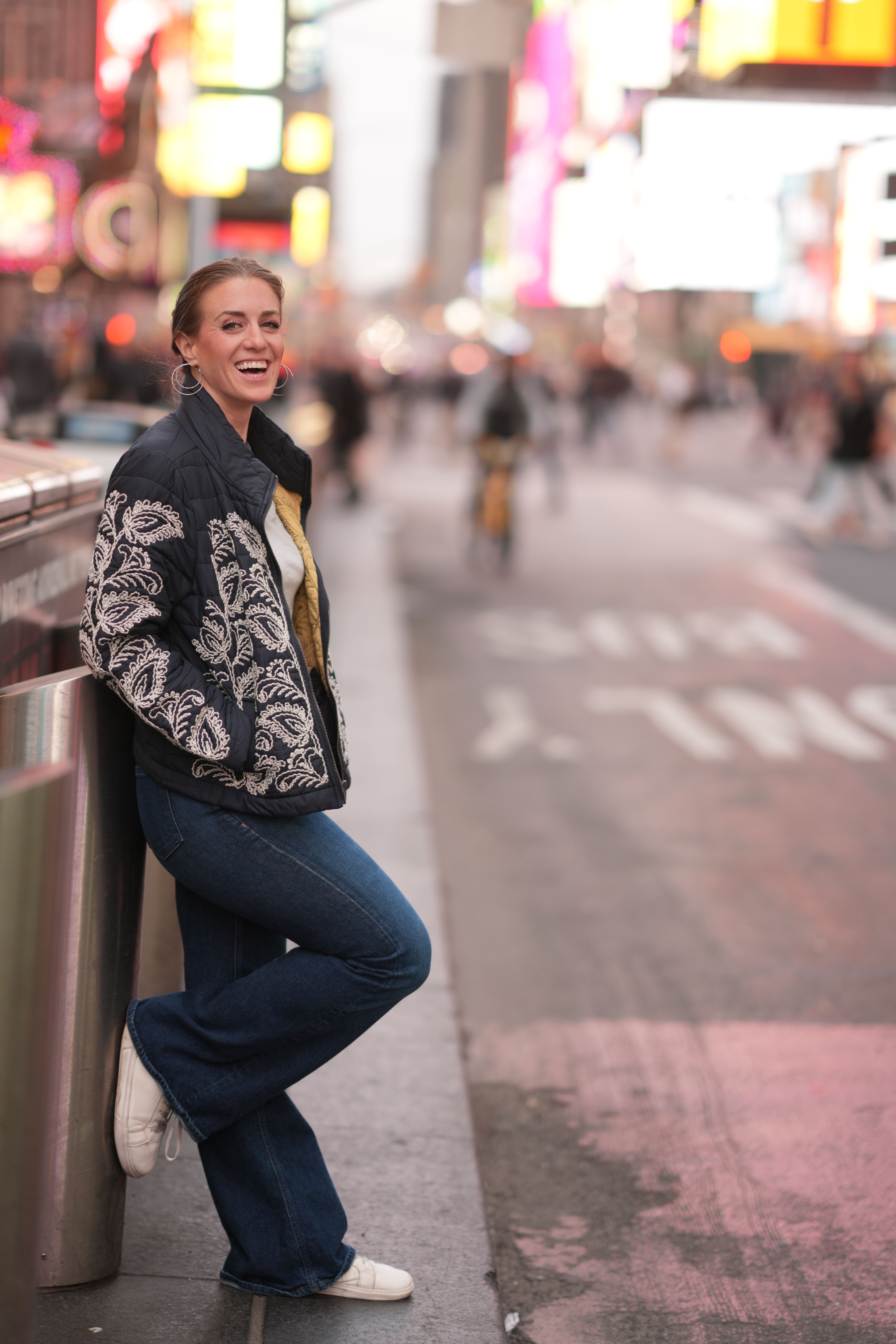 Woman leaning against a railing on a busy city street at night, illuminated by colorful lights.