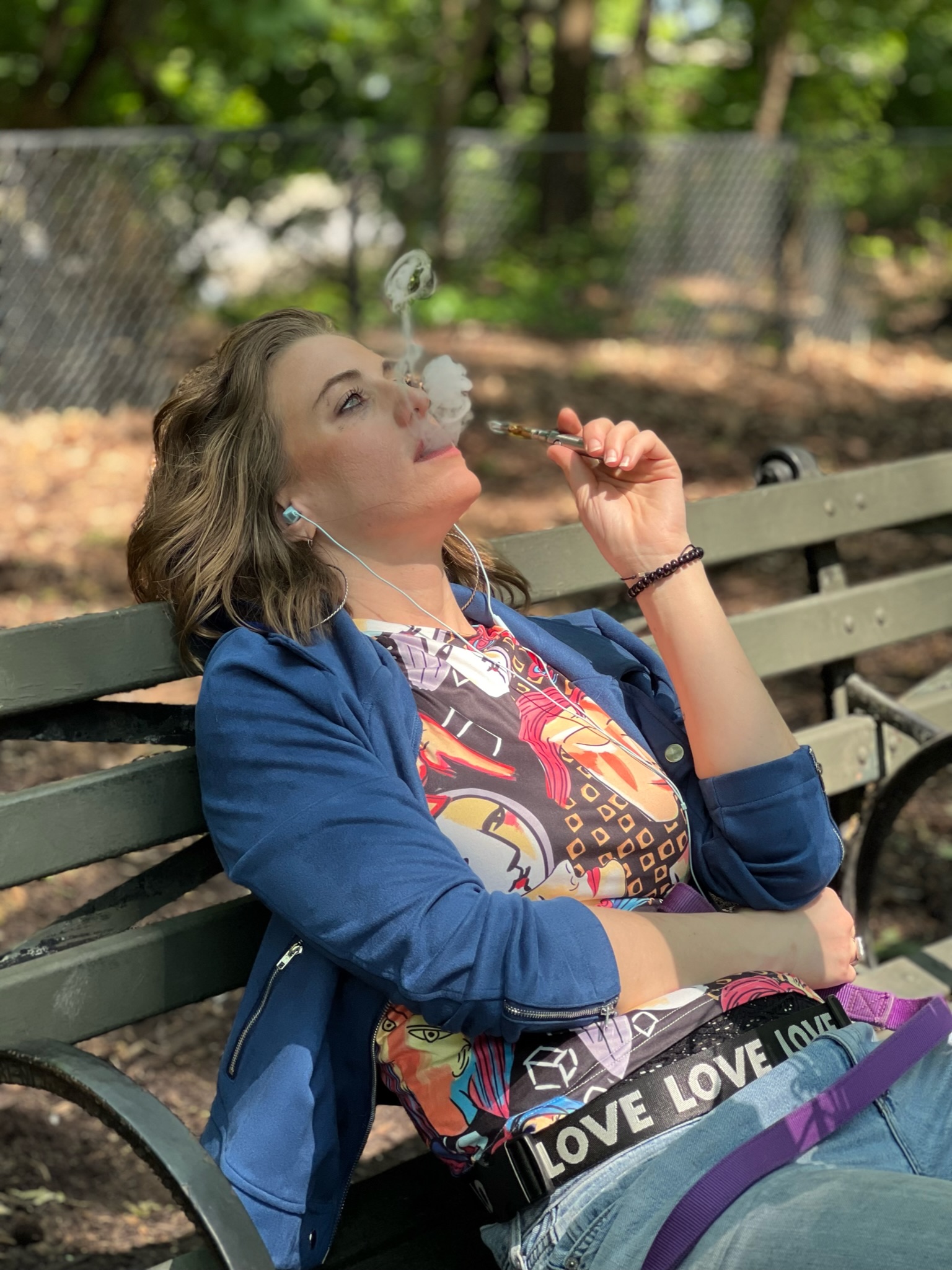 Woman sitting on a park bench blowing bubbles, surrounded by trees and sunlight, wearing a colorful shirt and blue jacket.