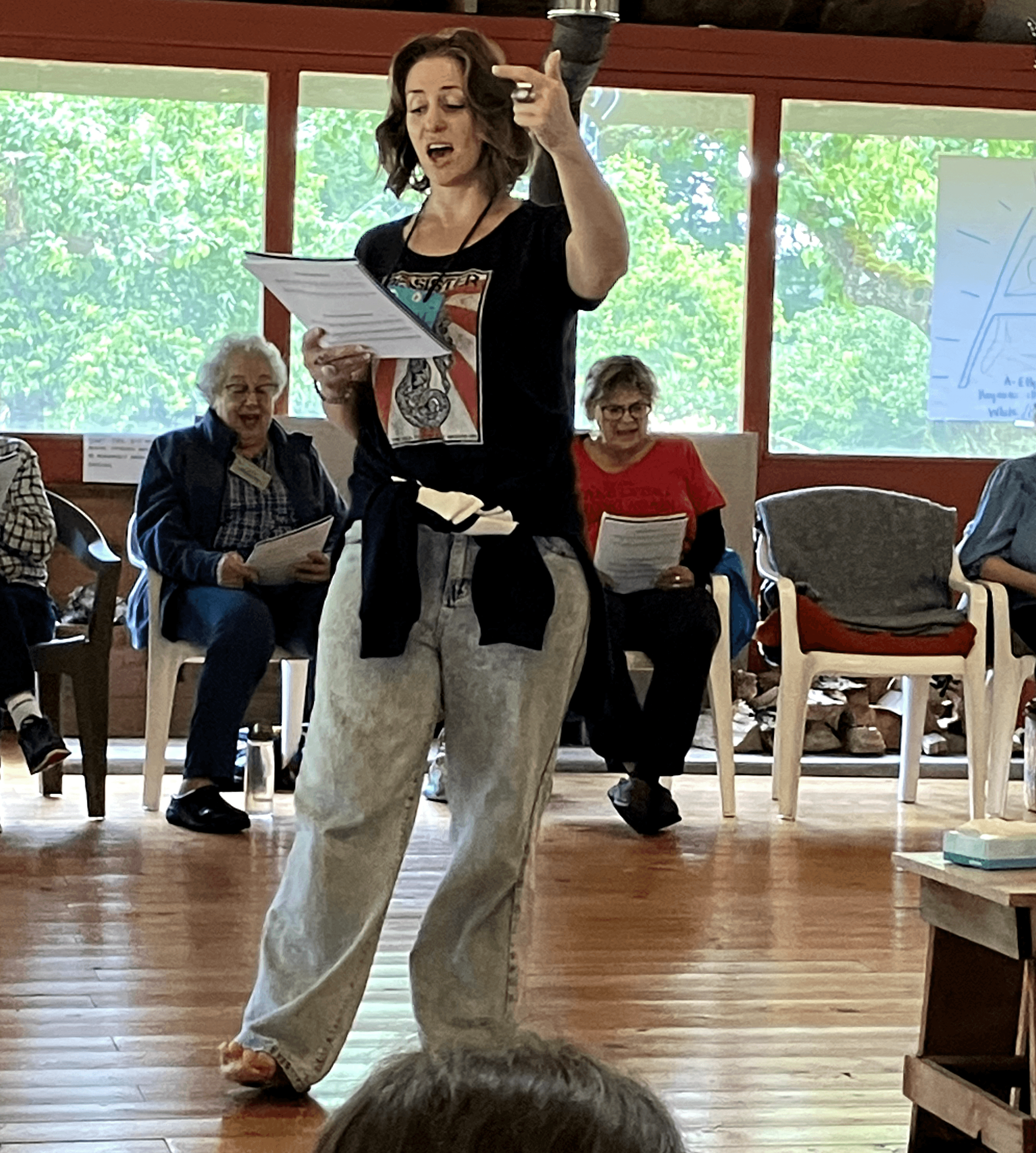 Woman standing and singing with a microphone, holding sheet music, in a room with seated people and large windows behind.