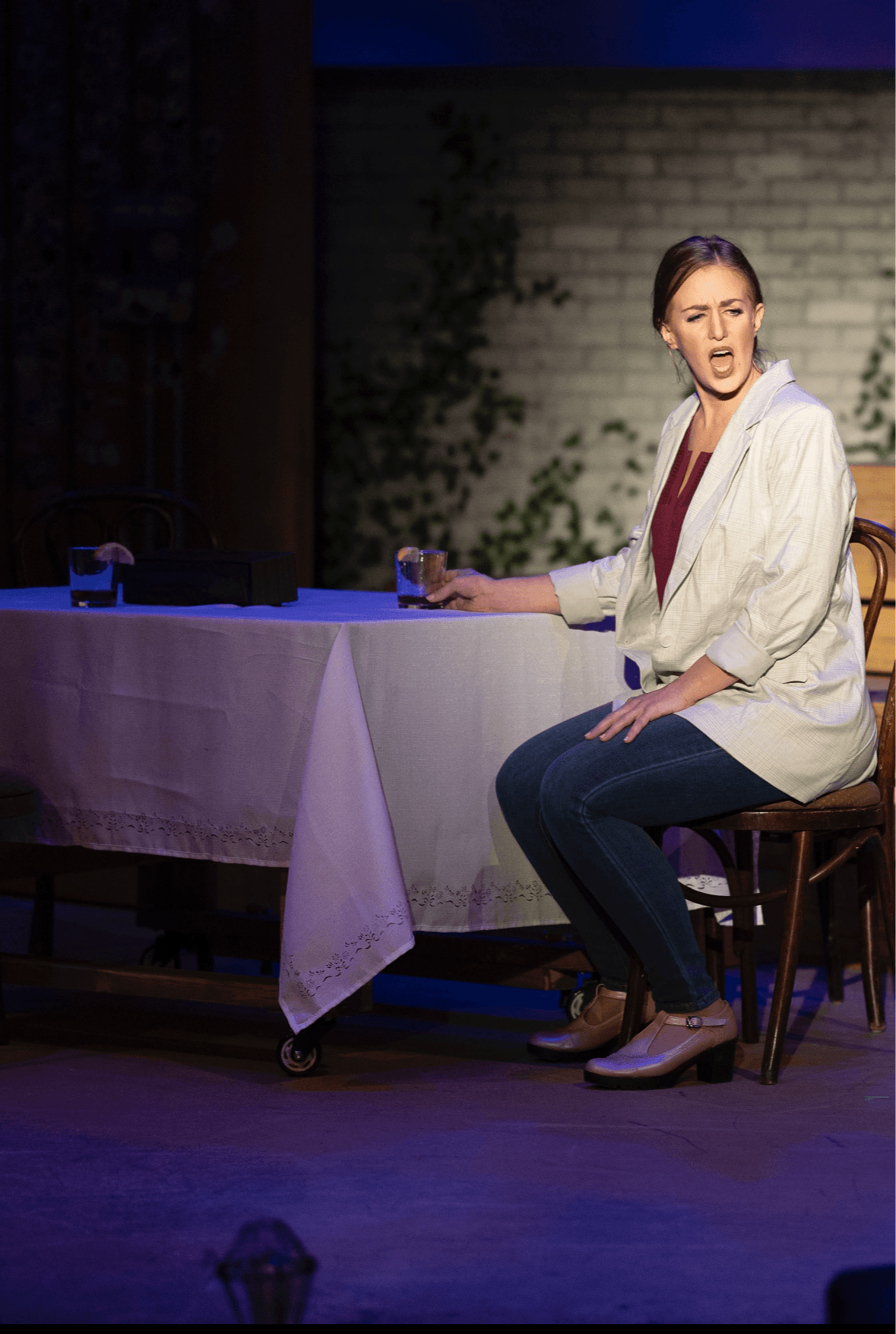 Woman sitting on a chair at a table, speaking or singing, with a brick wall background.