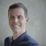 Portrait of a smiling man with short dark hair, wearing a dark collared shirt, against a plain background.