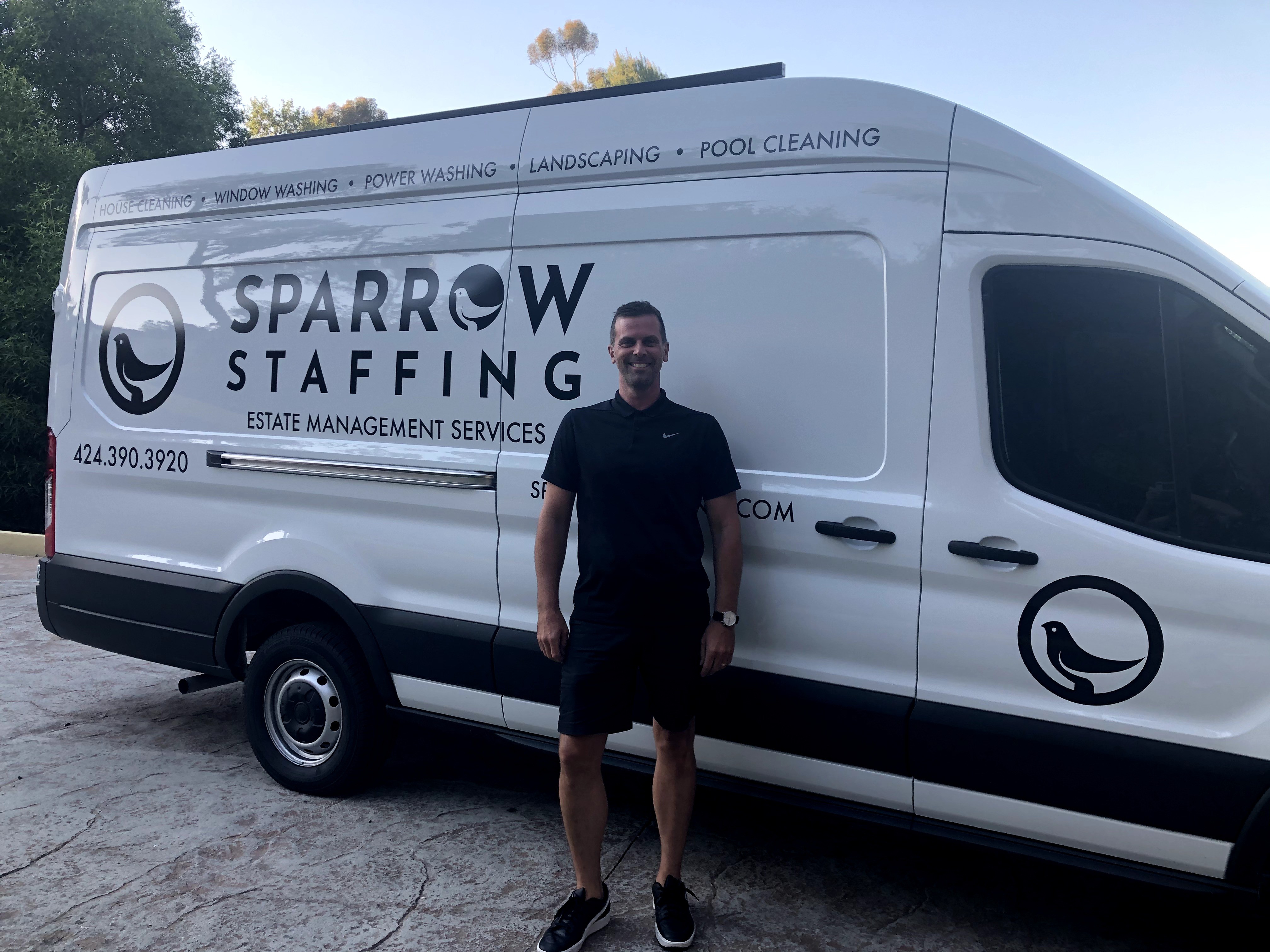 Person standing in front of a white van with company logo and text, outdoors on a paved surface.