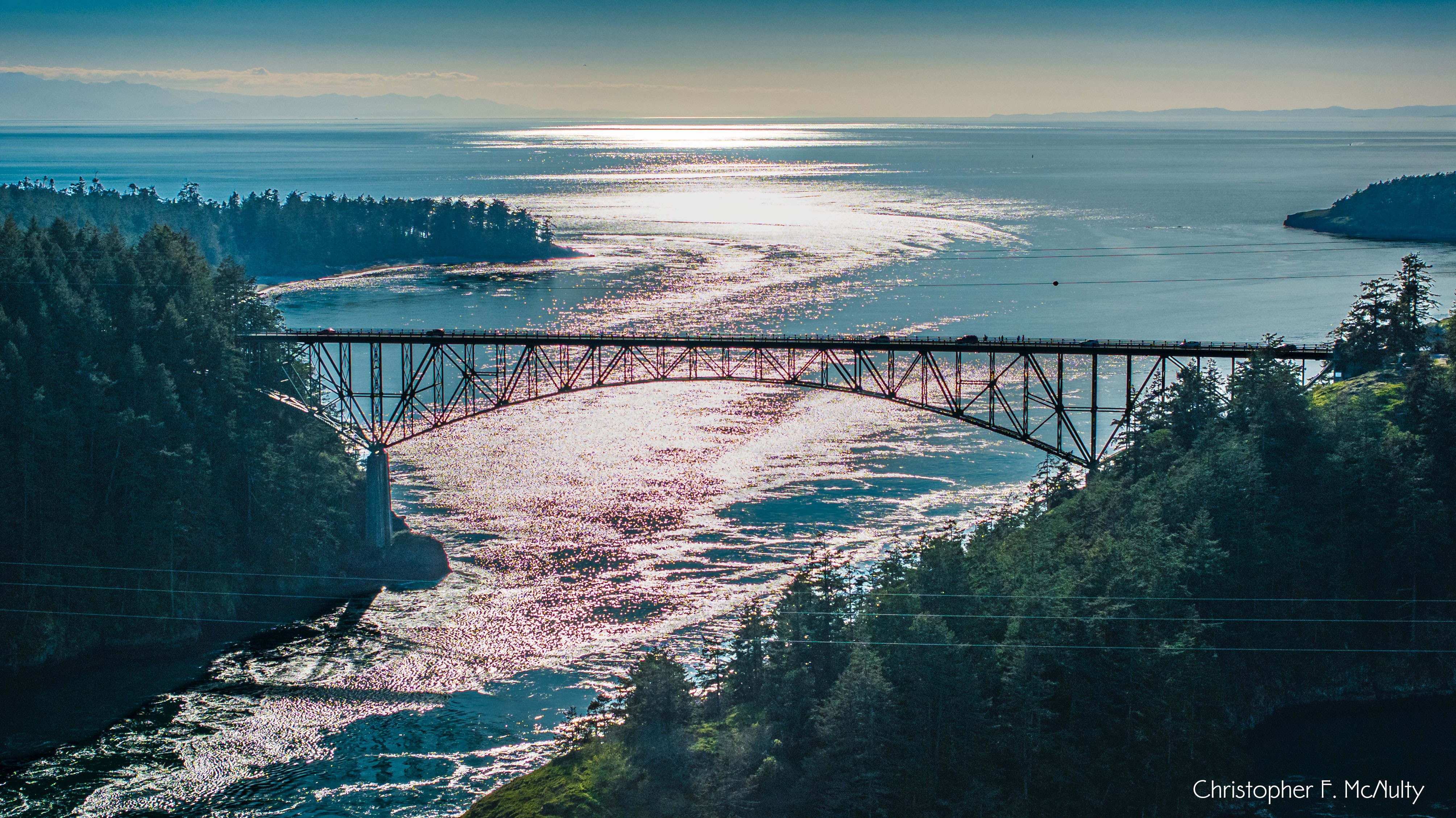 Bridge over a river with sunlight reflecting on water, surrounded by trees and distant land, under a clear sky.